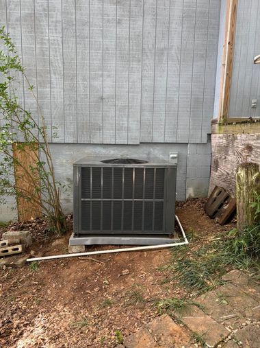 Outdoor HVAC unit against a gray building wall, surrounded by dirt and sparse plants.