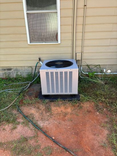 Outdoor air conditioner unit against tan house wall with a window above and red dirt ground below