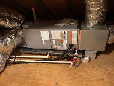 HVAC furnace and ductwork in an attic on a wooden floor with exposed pipes and vents