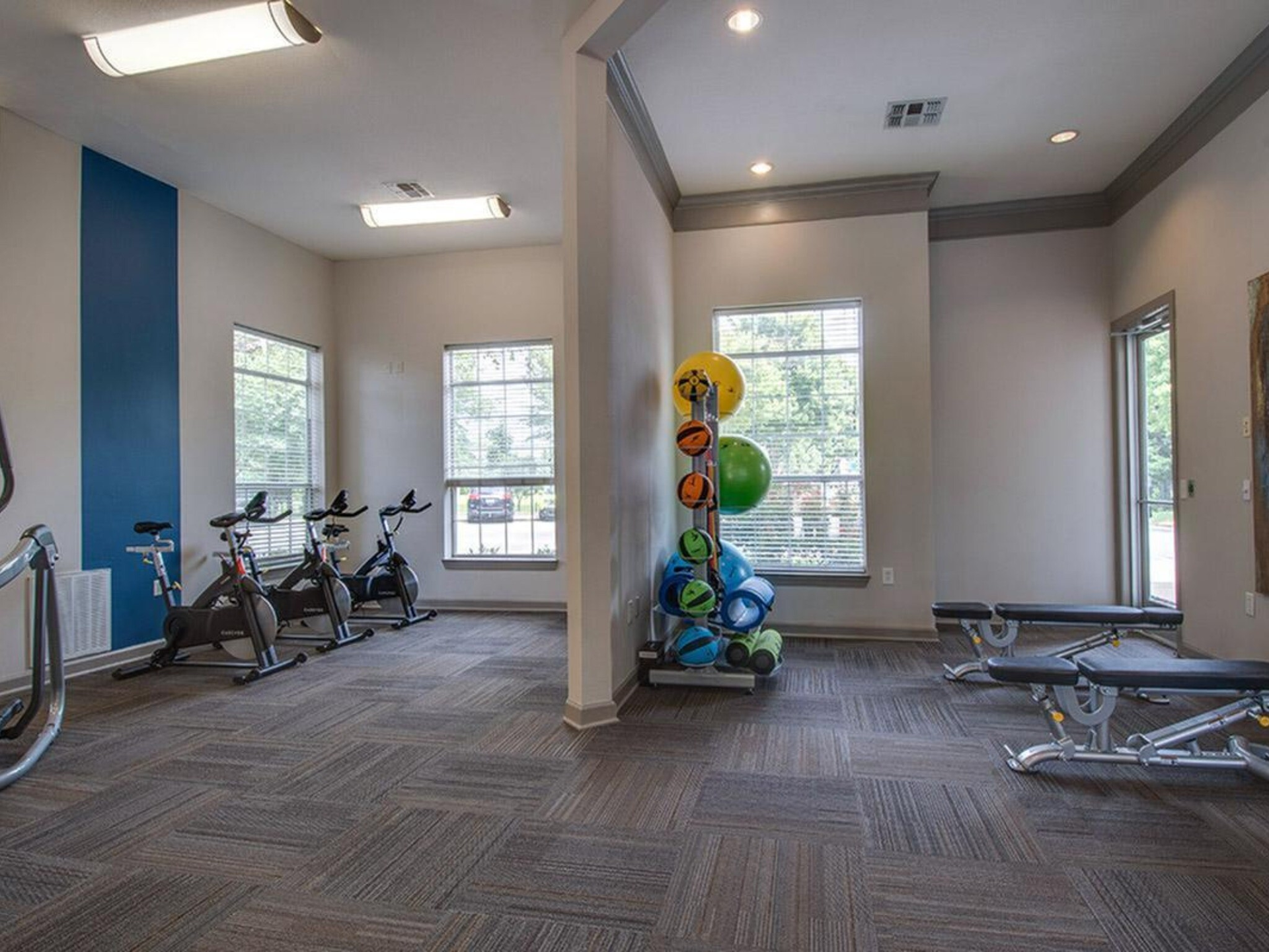 Gym with exercise bikes, weights, and medicine balls. Gray carpet, blue accent wall, and natural light from windows at Scout Landing.