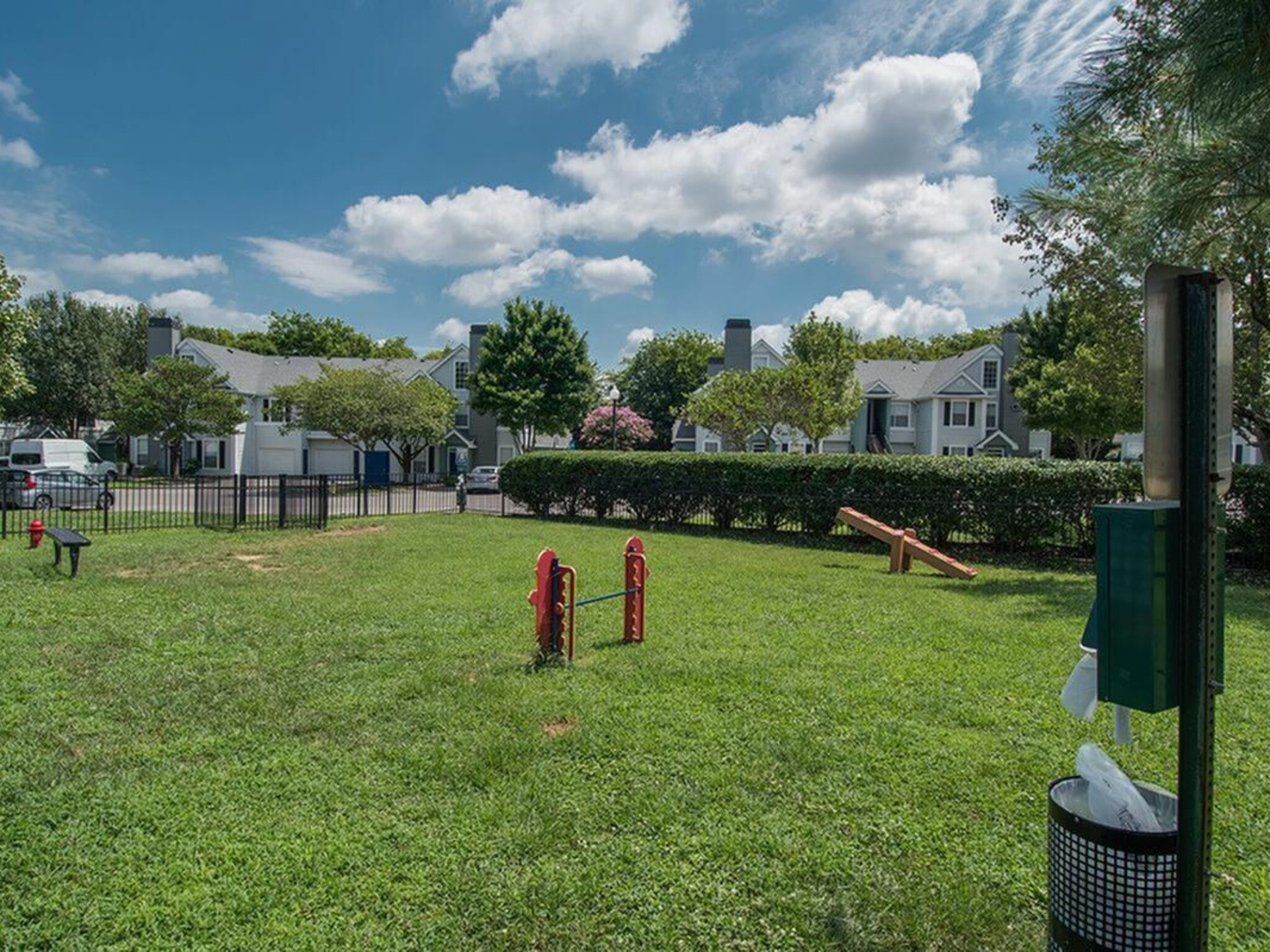 Dog park with green grass, agility equipment, and a waste bag dispenser. Apartment buildings are in the background under a cloudy sky at Scout Landing.