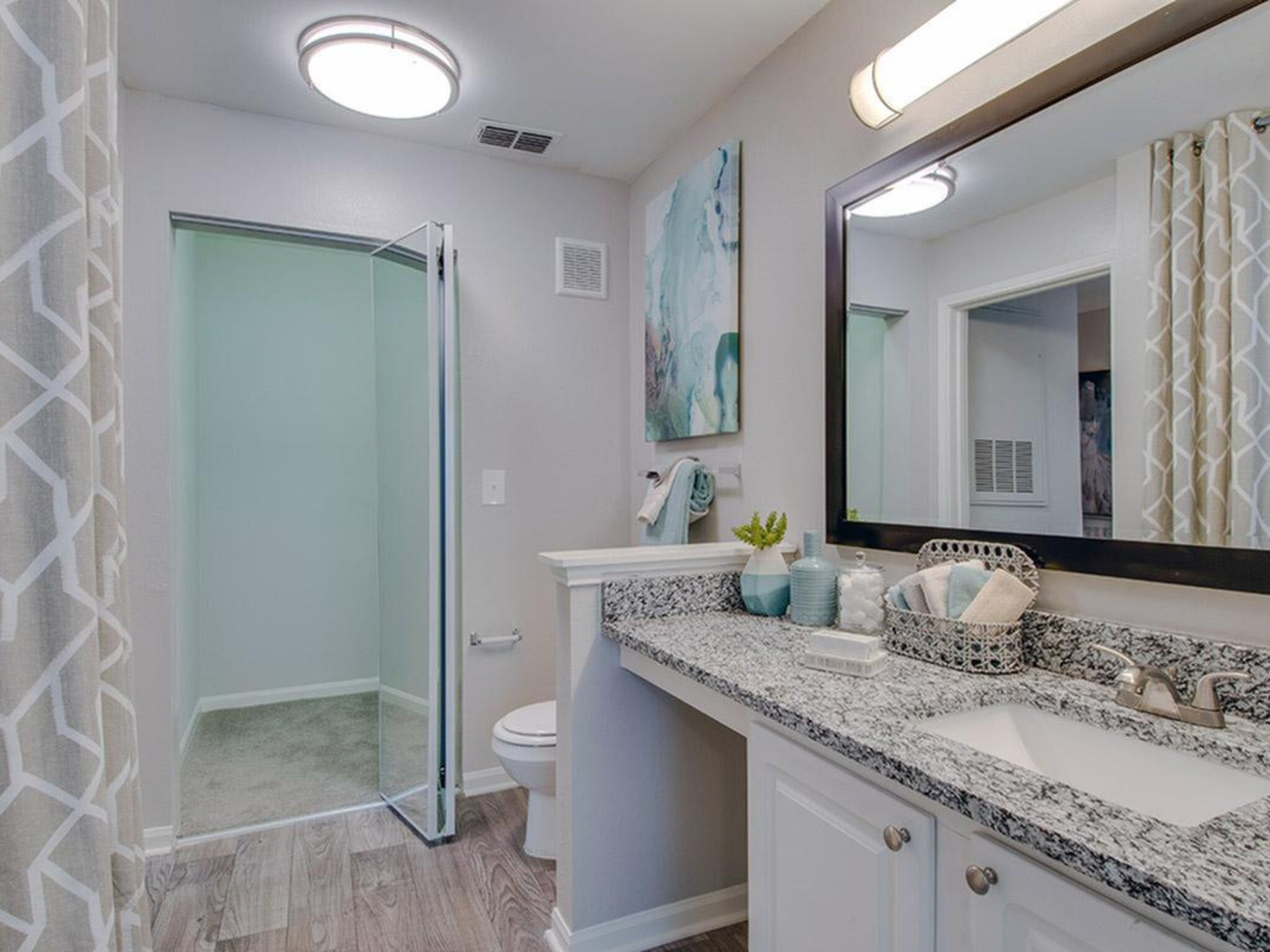 Bathroom with white vanity, granite countertop, large mirror, and glass shower. Soft color scheme with light gray walls and wood-look floor at Scout Landing.