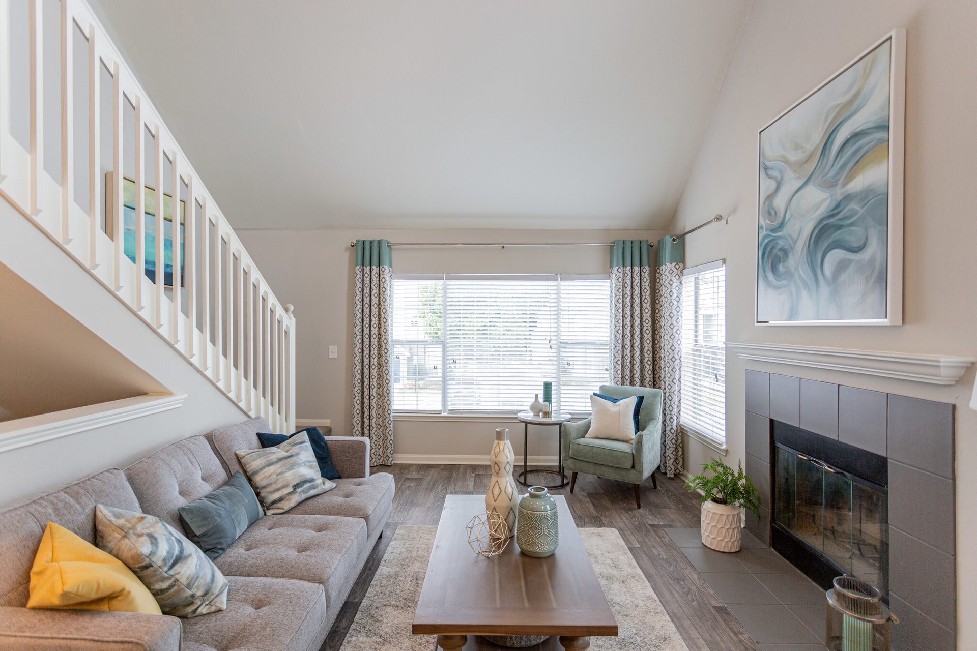 Living room with a grey sofa, a fireplace, and a staircase.  A large window in the center with patterned curtains at Scout Landing.