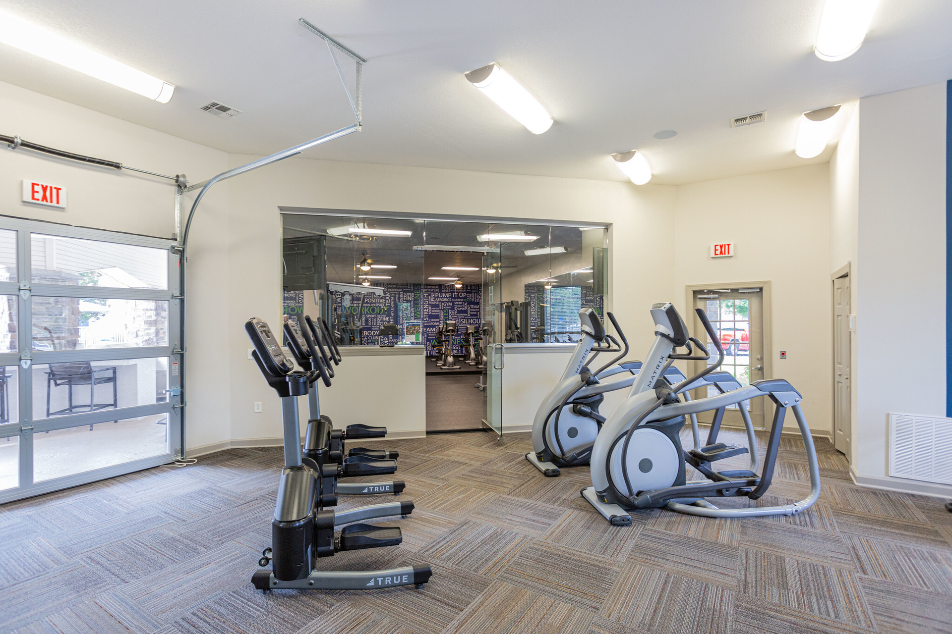 A fitness room with exercise bikes and elliptical machines.  A large glass door and window offer an outside view at Scout Landing.