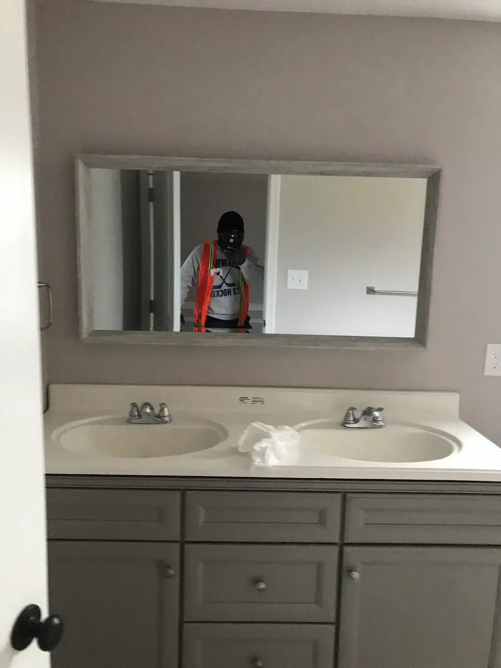 Bathroom with double sinks, gray vanity, mirror reflecting a person in orange vest.