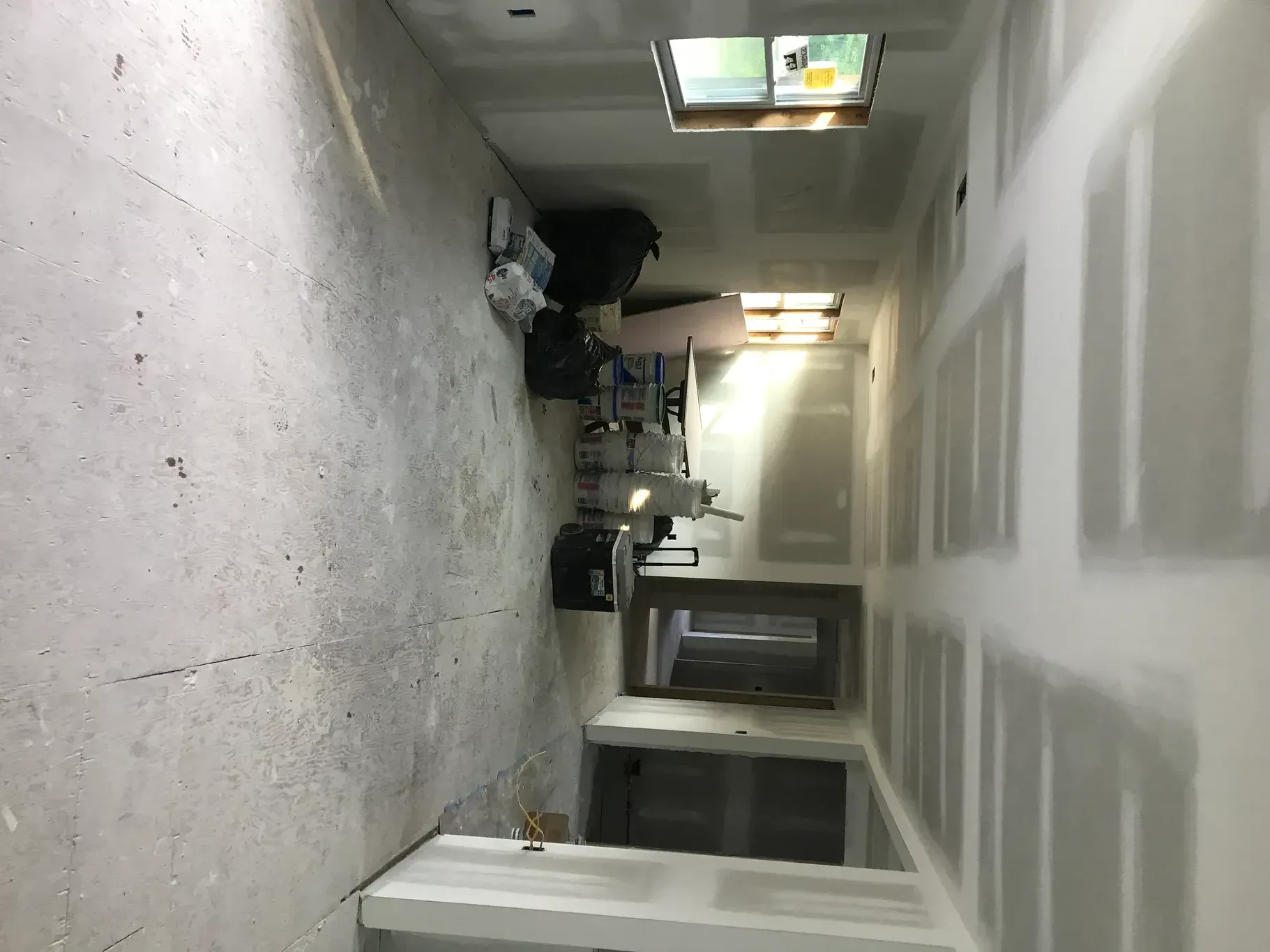 A long, unfinished room with drywall, tools, and a window.