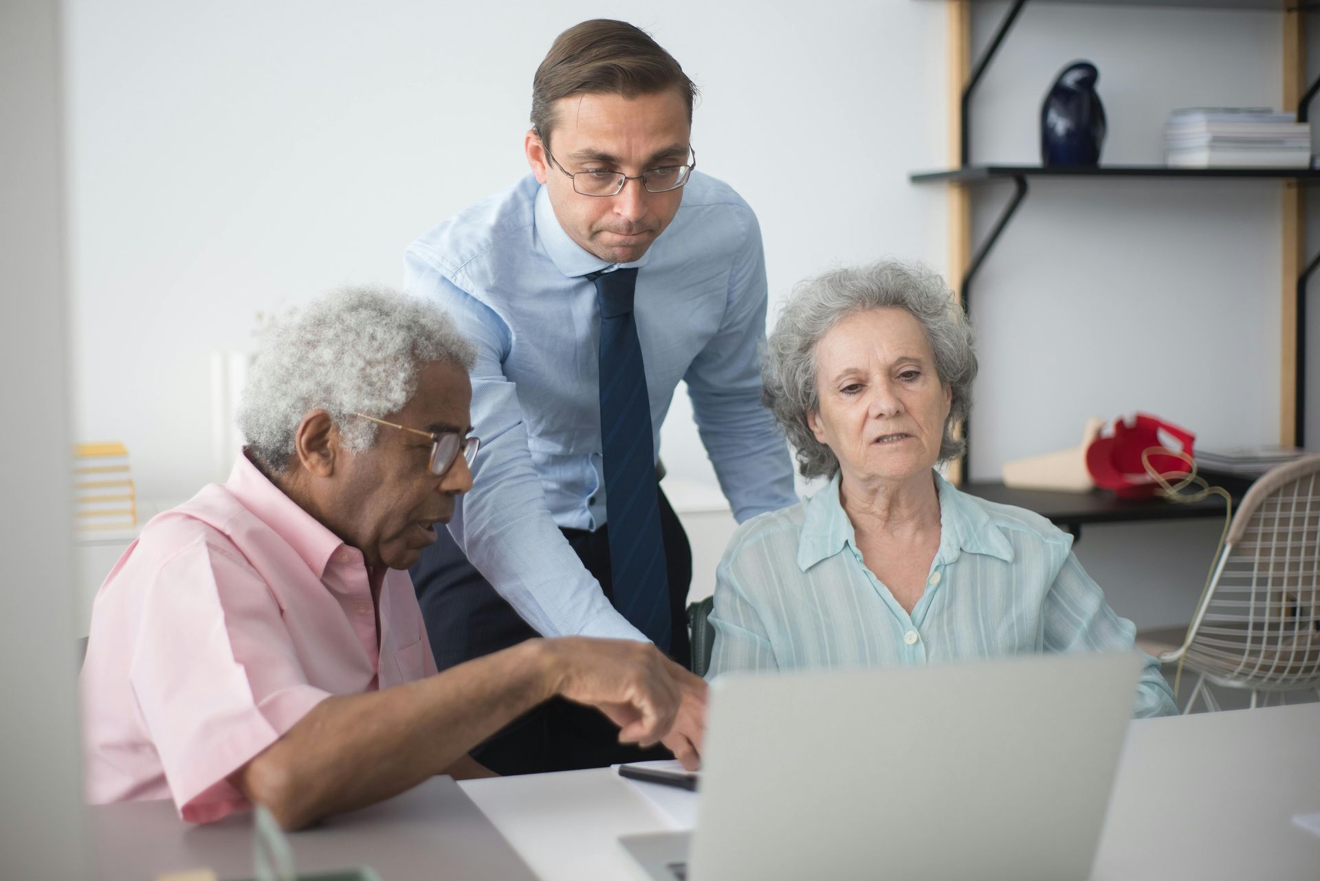 A young man in a suit helps two older adults use a laptop in an office setting.