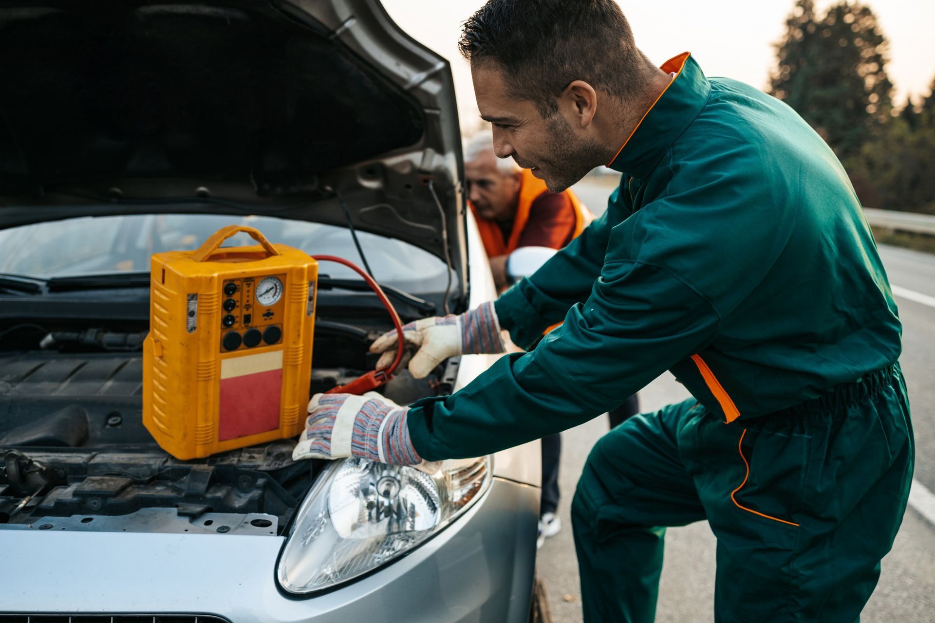 Mechanic jump-starts a car on the roadside using a yellow portable jump starter; older person watches.