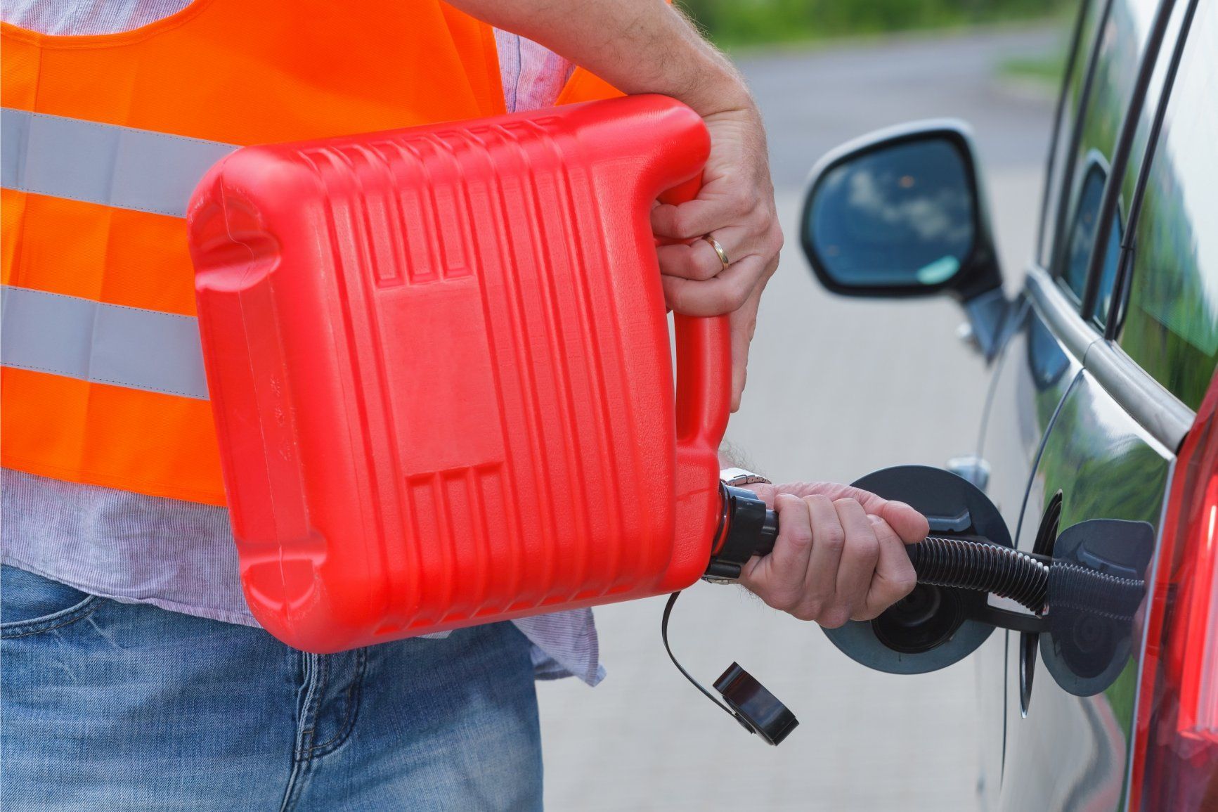 Person filling car with gas from a red gas can, wearing an orange safety vest, roadside.