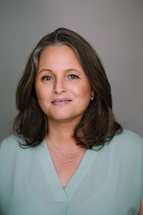 Frau mit braunem Haar, hellgrünes Oberteil tragend, lächelt vor grauem Hintergrund in die Kamera.