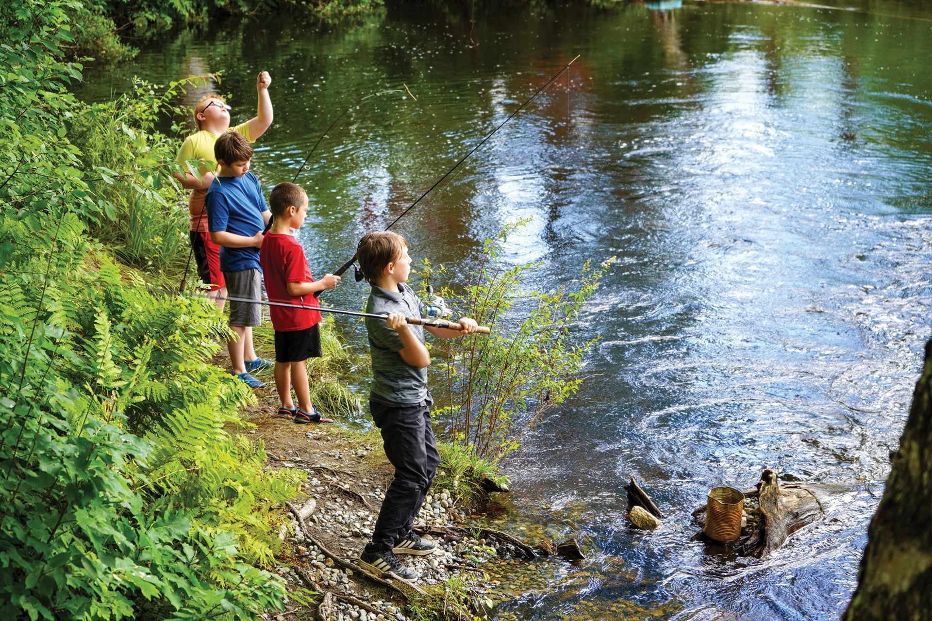 Four children fishing with rods on the bank of a wooded stream.