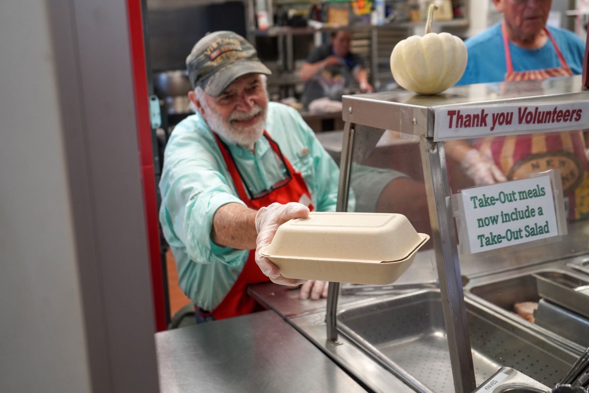 Volunteer hands a take-out meal, smiling. Red apron, light green shirt. Counter setting with “Thank you Volunteers” sign.