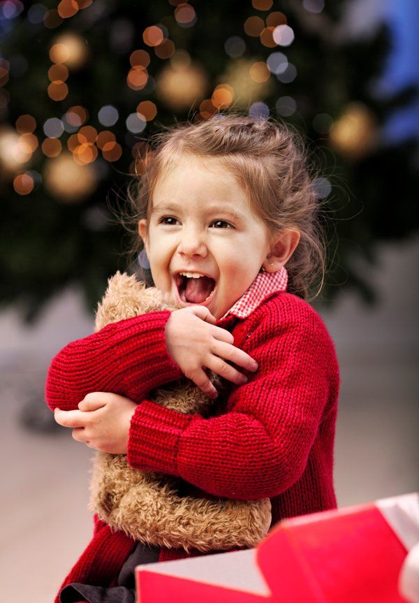 A little girl is holding a teddy bear in front of a christmas tree.