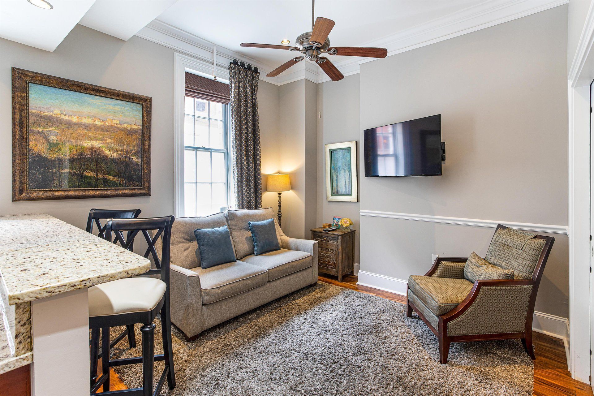 Cozy living room with sofa, armchair, rug, mounted TV, and artwork. Includes a bar and a ceiling fan.