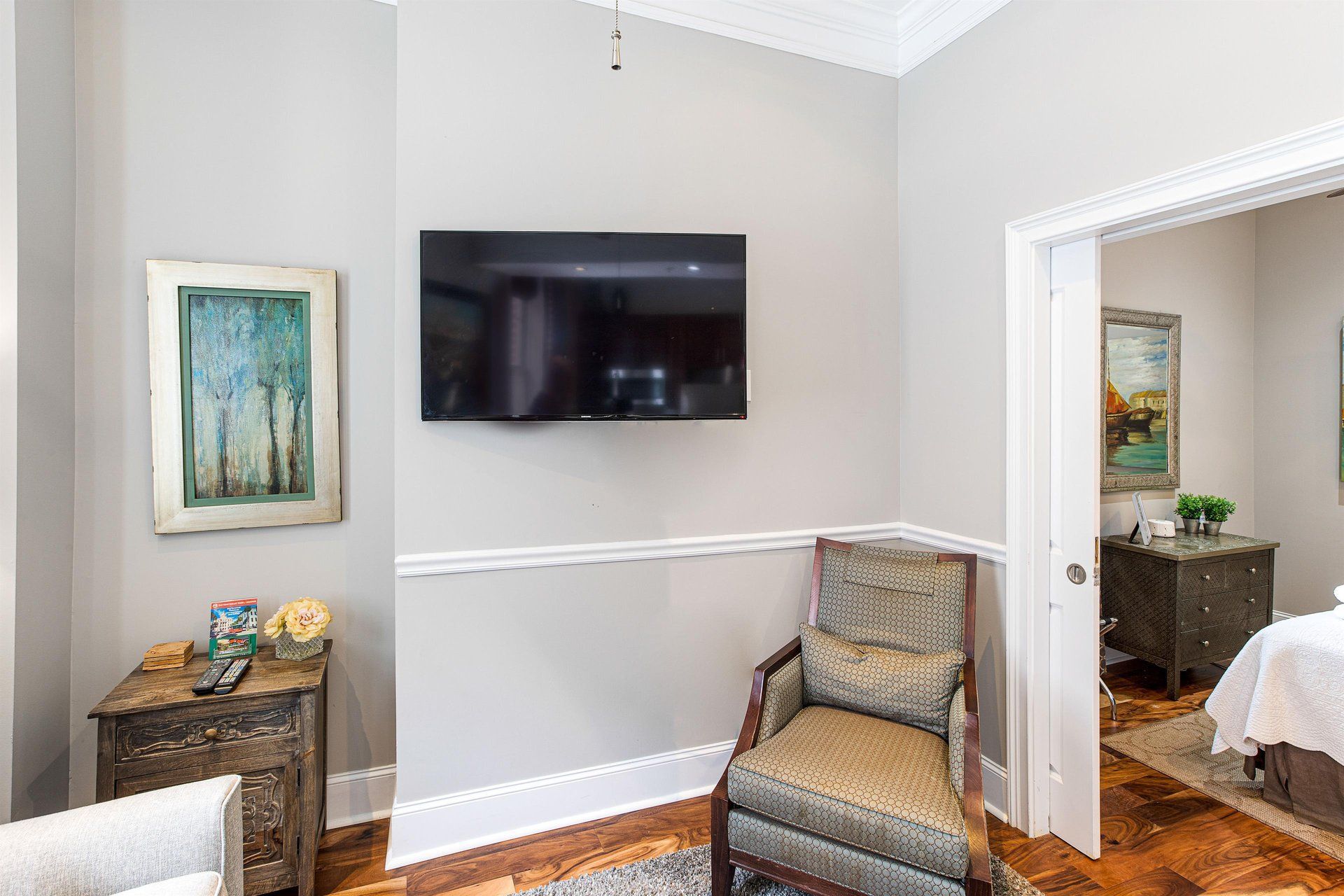 A living room with a TV, a chair, and a painting; a doorway leads to a bedroom.