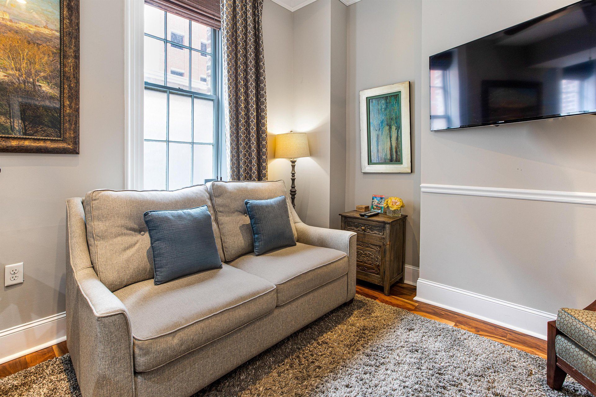Living room with couch, TV, window, art, and rug. Gray walls and wood floor.
