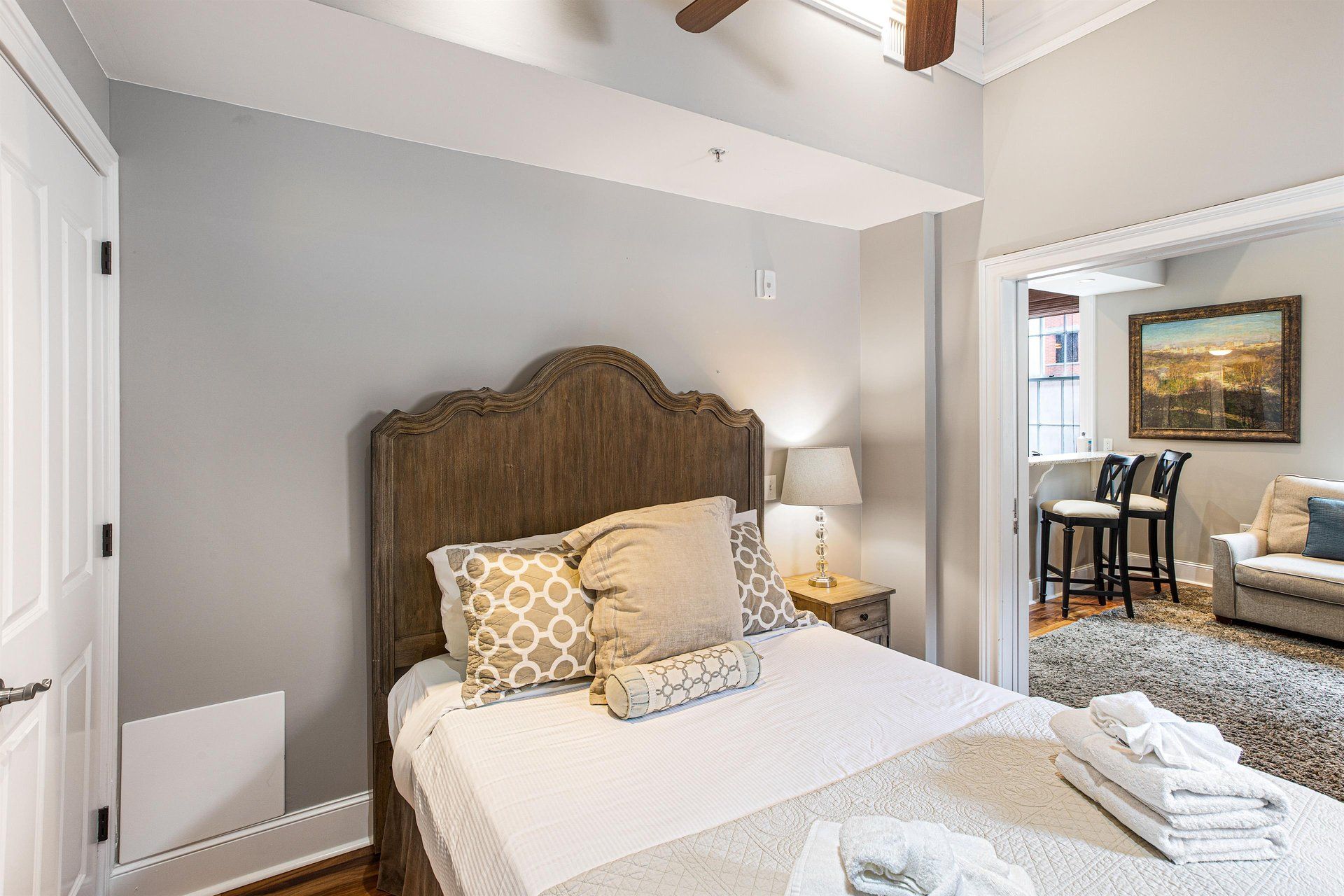 Bedroom with wooden headboard, soft bedding, and an opening to a living area with artwork and furniture.