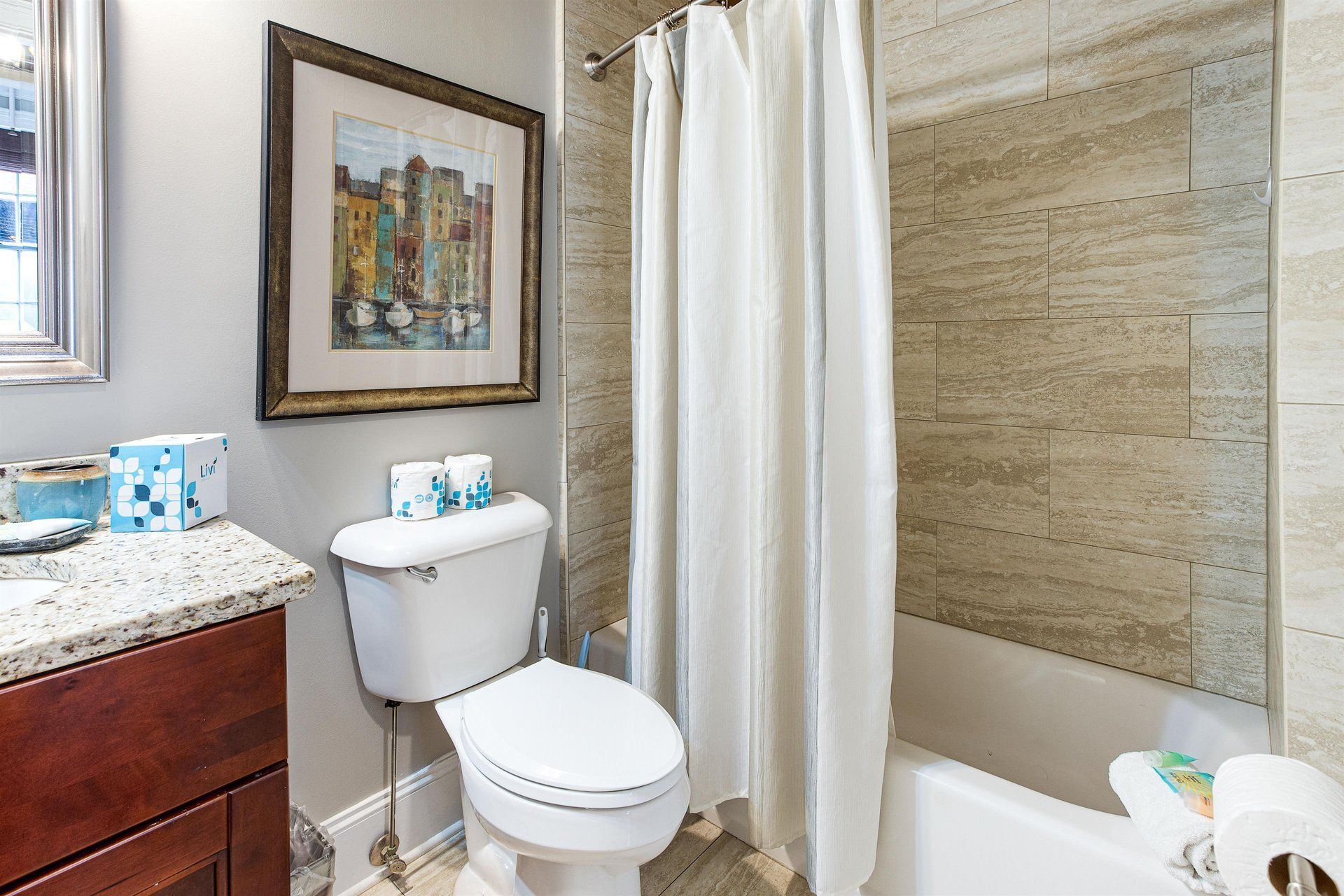 Bathroom with a white toilet, shower, and vanity. Beige tile wall and artwork on the wall.