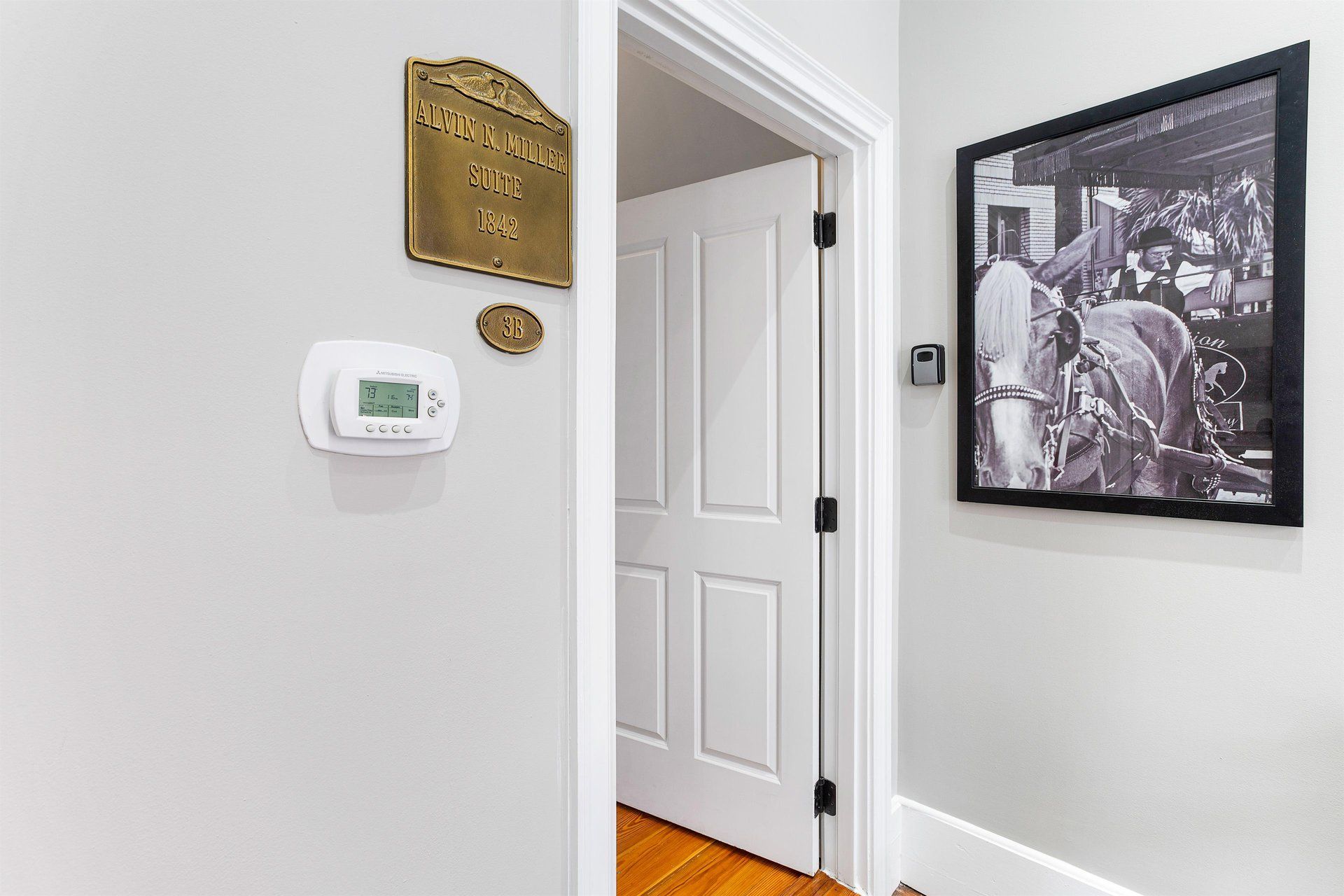 Hallway with white walls, a thermostat, framed photo, and a partially open door.