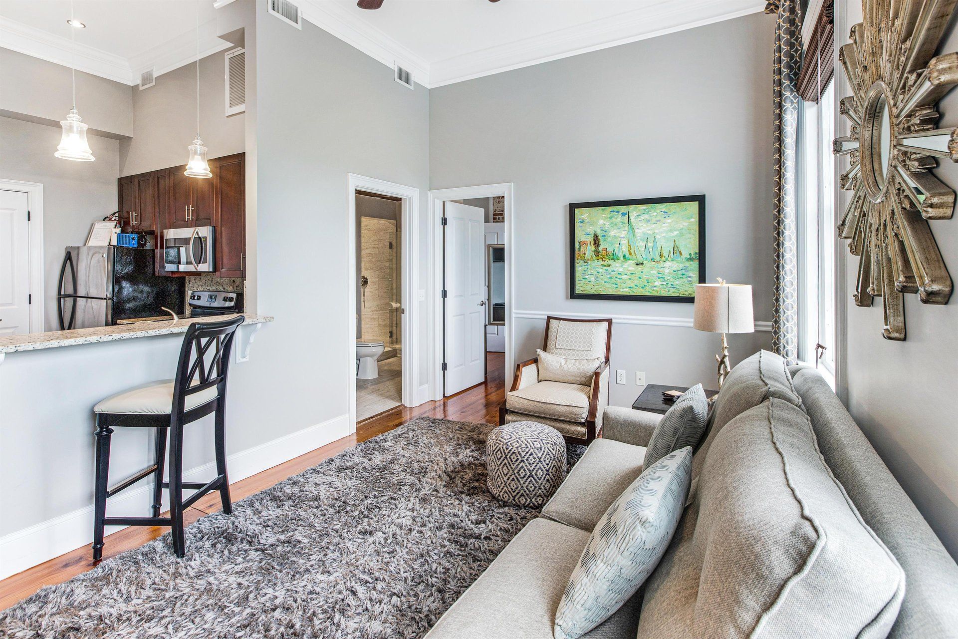Living area with kitchen, couch, armchair, rug, artwork, and sunlight through window.