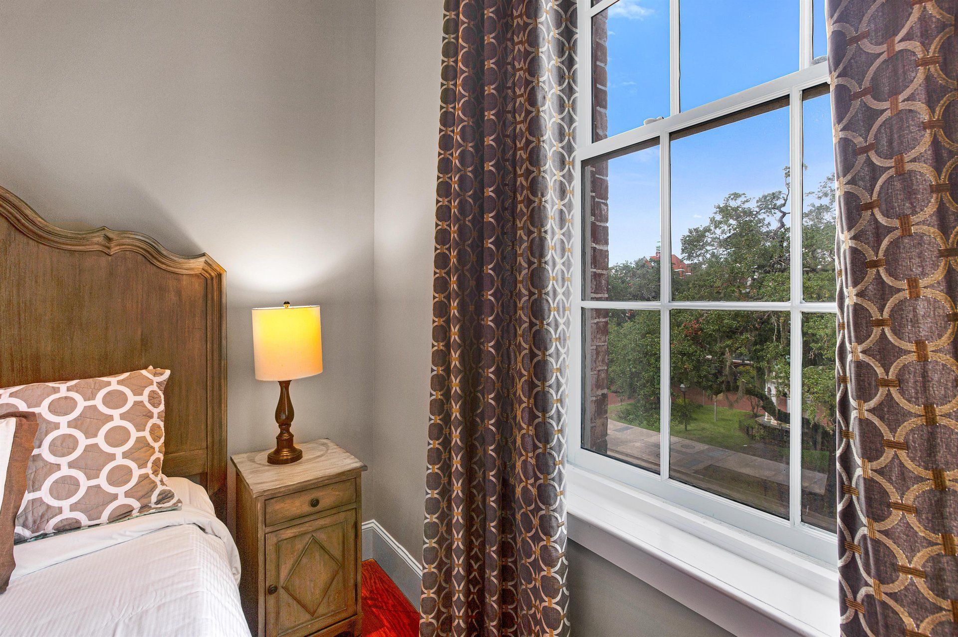 Bedroom with wood headboard, bedside table with lamp, patterned curtains, and window overlooking a green area.
