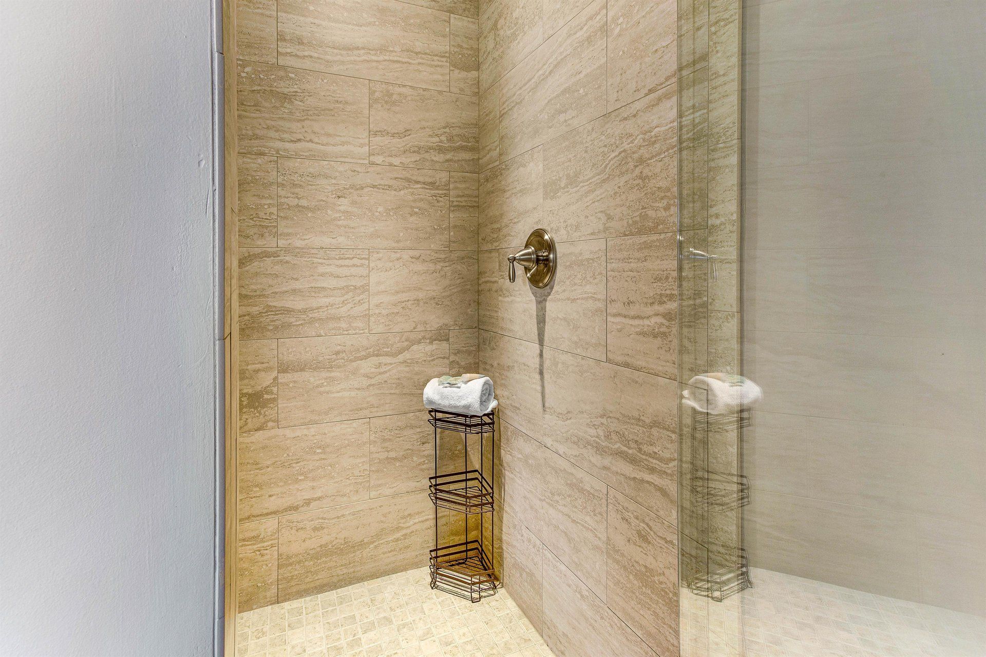 Shower stall with beige tile walls, glass door, and chrome fixtures. A small shelf holds towels.