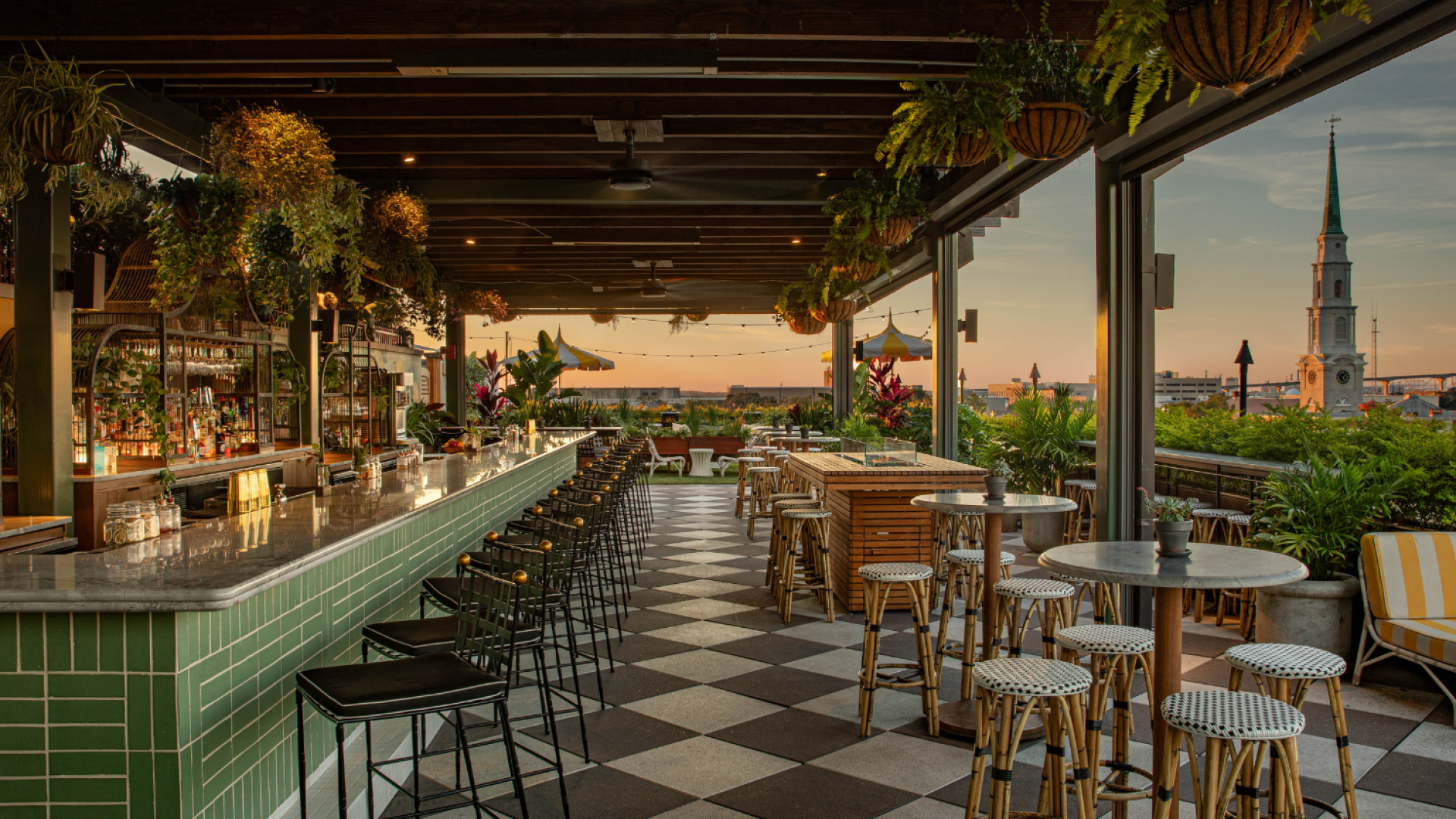 Rooftop bar with a tiled floor, counter, and stools, overlooking a cityscape at sunset. Plants hang overhead.