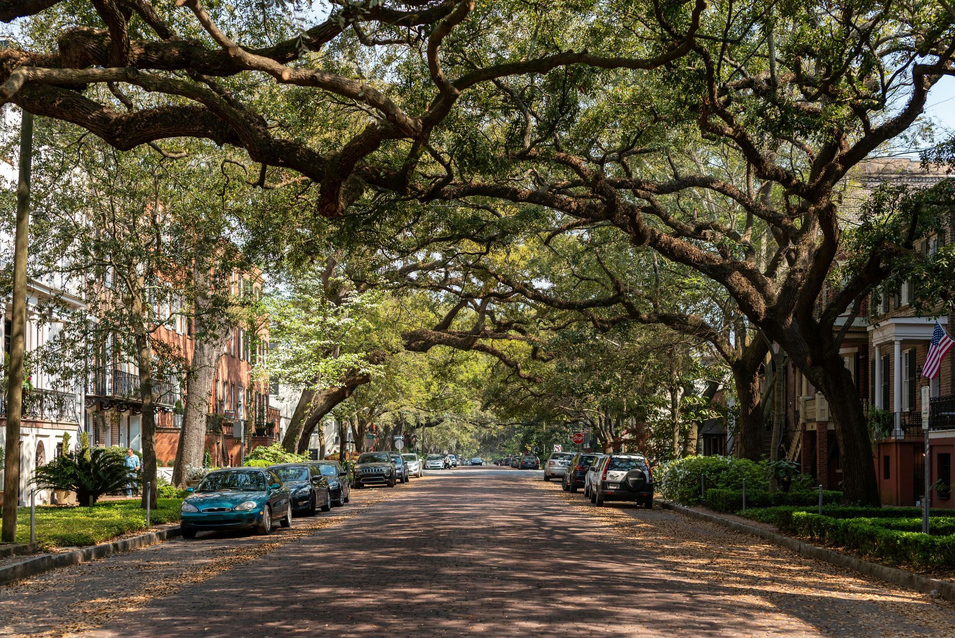 Street lined with trees, cars parked, brick road, historic buildings with porches in Savannah, Georgia.
