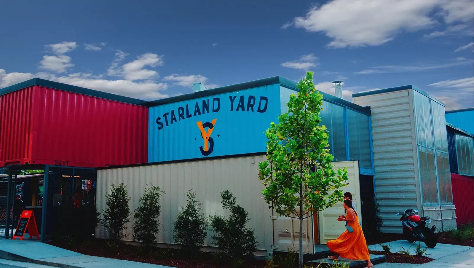 Starland Yard, colorful building made of shipping containers; person in orange dress near a tree.