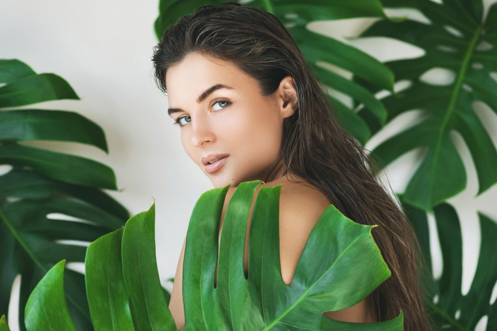 Woman with long brown hair, blue eyes, partially hidden by large green Monstera leaves.