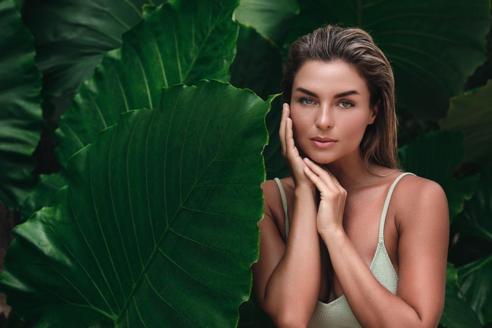 Woman in a white swimsuit poses in front of large, green leaves, touching her face, looking directly at the viewer.