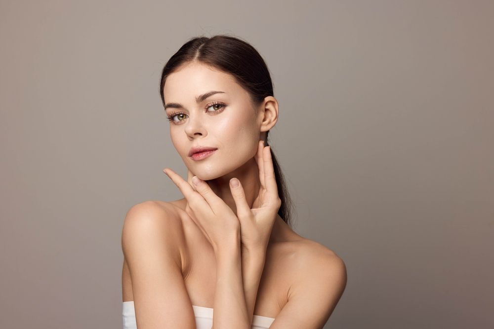 Woman with clear skin, hands near face, light background, looking at the viewer.