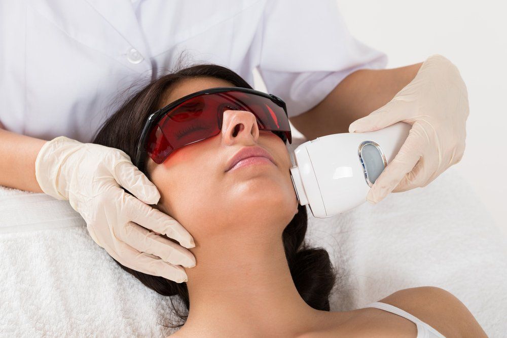 Woman undergoing laser facial treatment; esthetician holds device, patient wears protective eyewear.