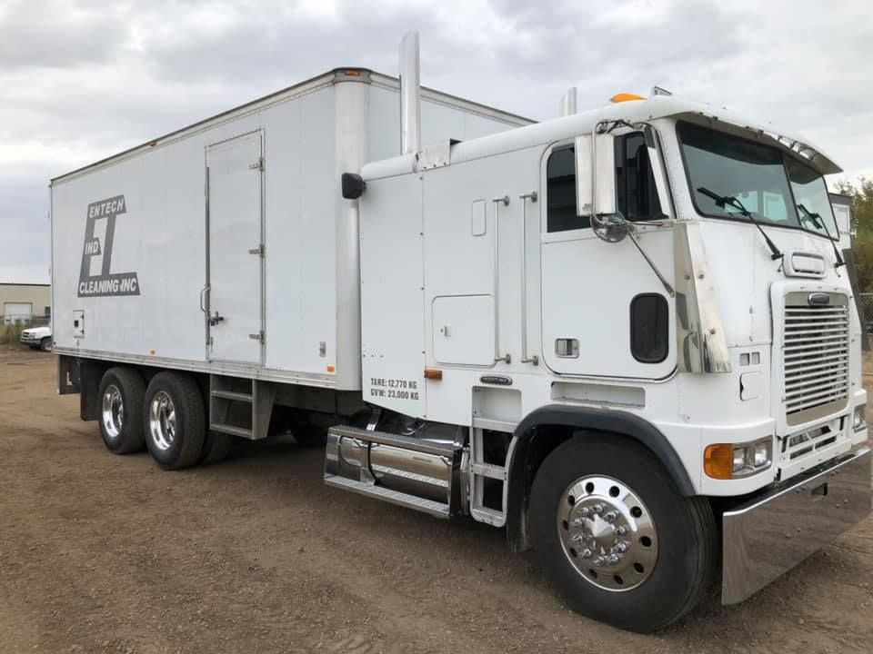 A white semi truck with a box on the back is parked in a dirt lot.