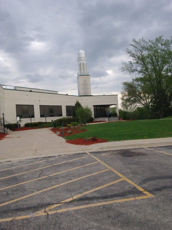 Church building with white steeple beside a parking lot and landscaped lawn under cloudy skies