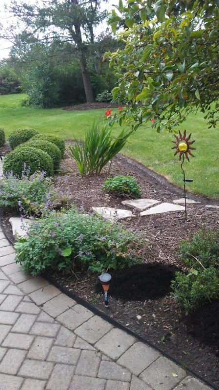 Backyard garden bed with shrubs, flowers, and decorative metal sun stakes beside a paved walkway