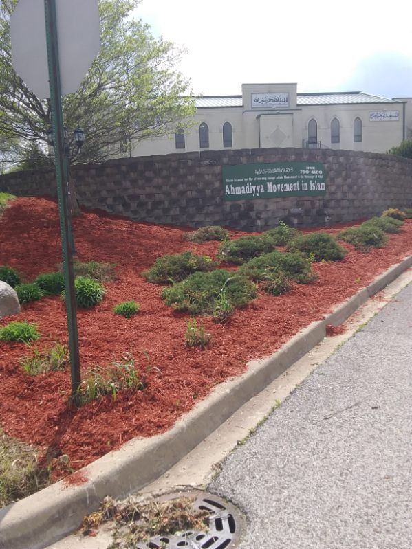 Landscaped entrance with red mulch, shrubs, stone wall sign, and a building in the background.