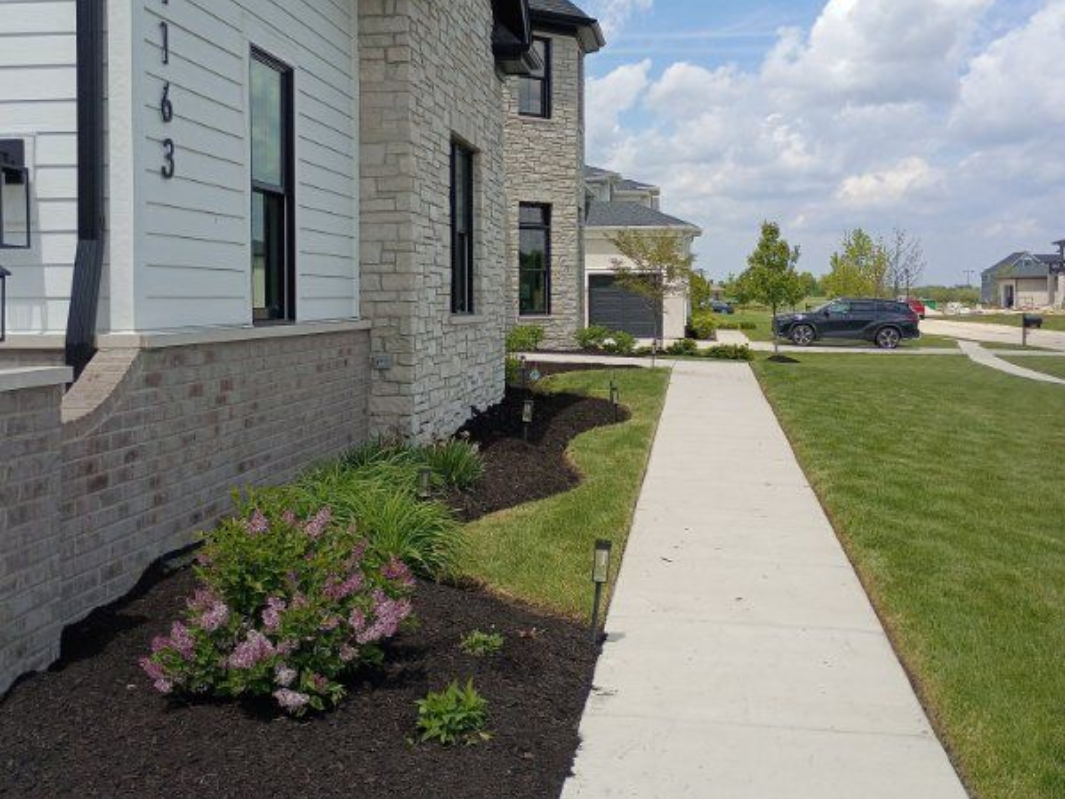 Sidewalk beside a stone house with mulched flower beds and a grassy lawn in a suburban neighborhood.
