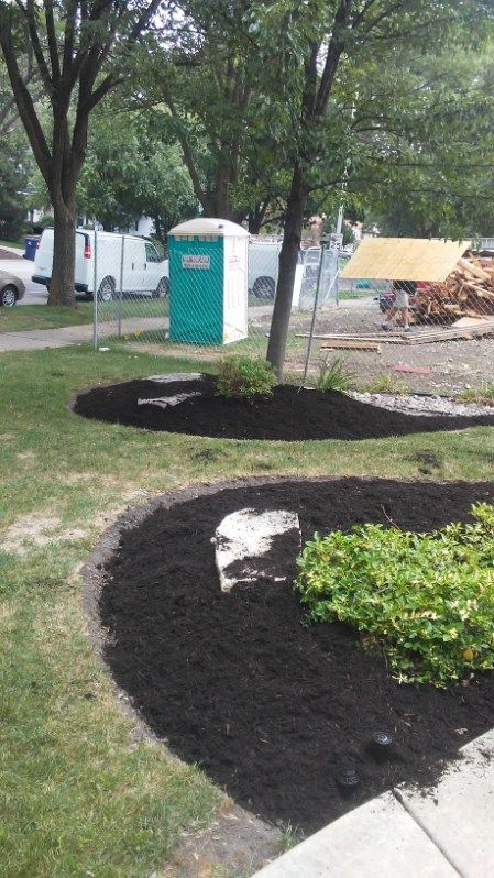 Freshly landscaped park bed with dark mulch, green shrubs, trees, and a portable toilet in the background