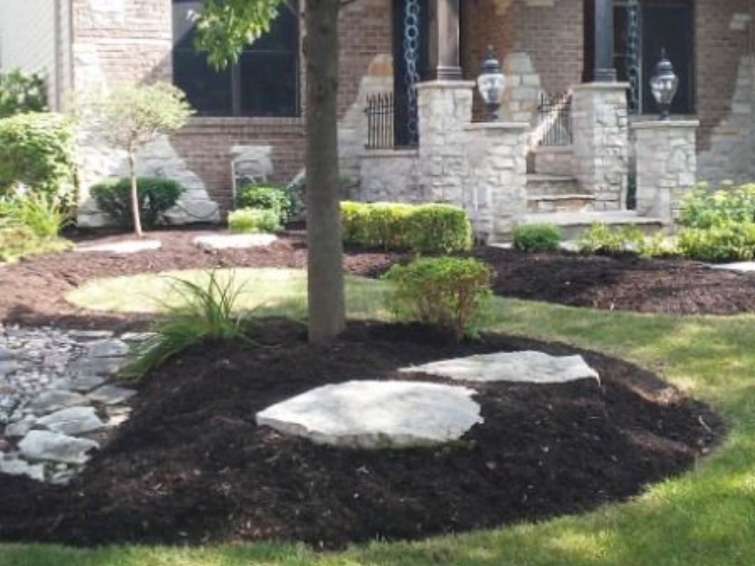 Front yard landscaping with a tree in a mulched bed, white rocks, and a stone house porch in the background