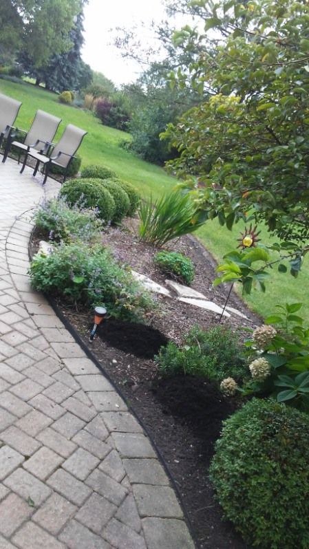 Curved stone patio beside a landscaped garden with chairs, shrubs, and green lawn