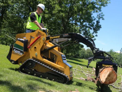 Worker using a yellow Vermeer tracked loader to cut a tree trunk in a grassy area.