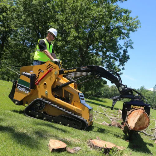 Worker operating a yellow Vermeer compact loader moving a large tree log in a sunny grassy field