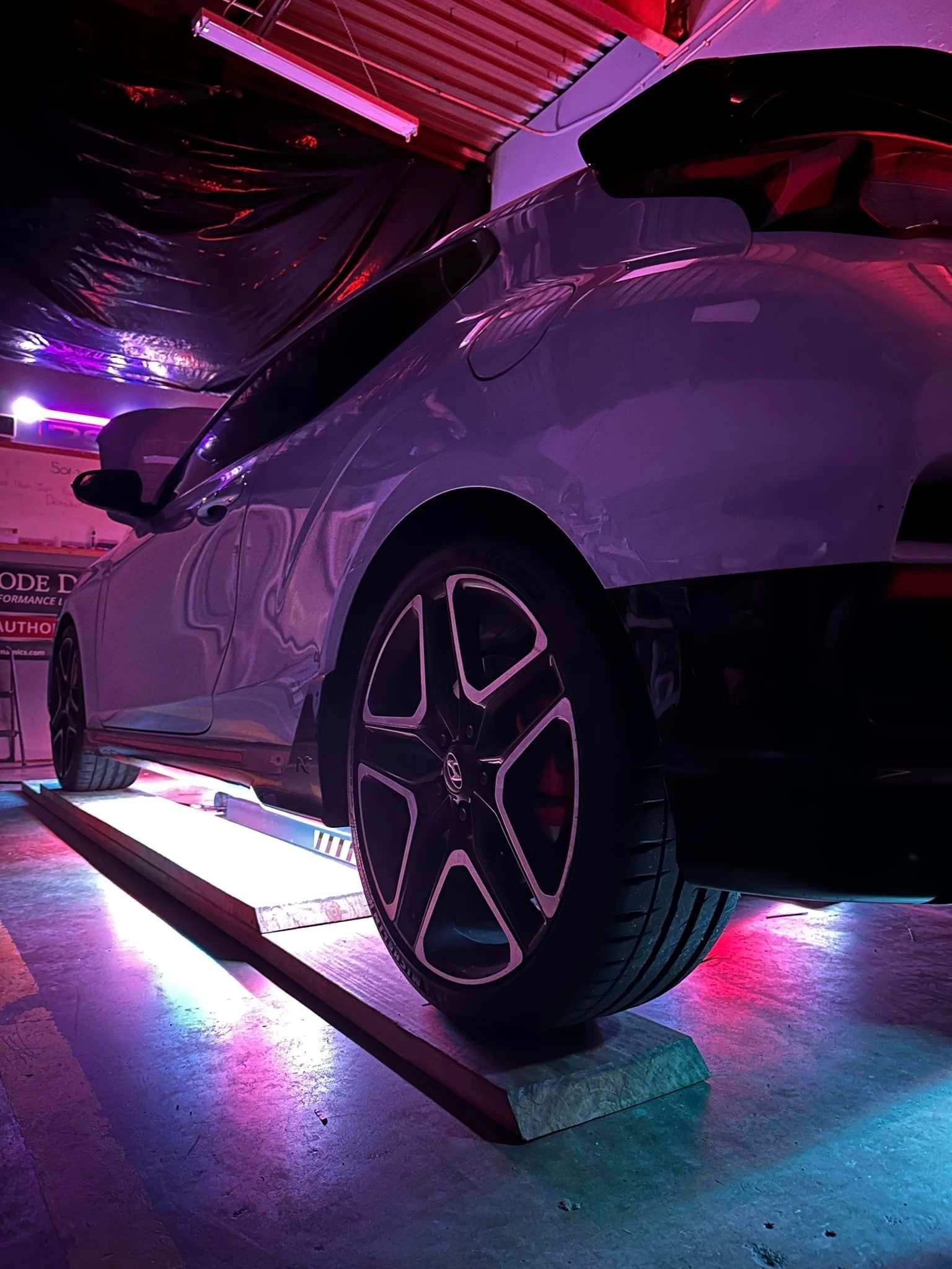 White car with glowing neon underlights in a purple-lit garage, rear wheel visible