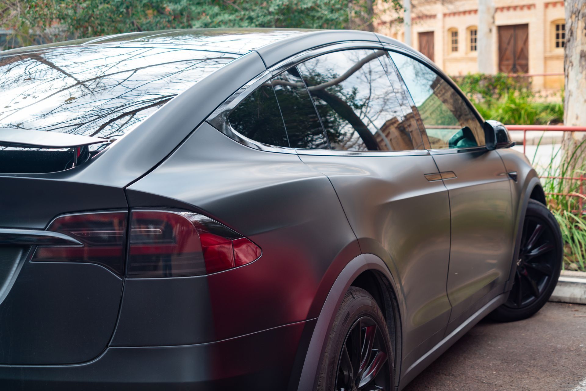 Black Tesla Model X parked on a street beside a building with trees reflected in the windows
