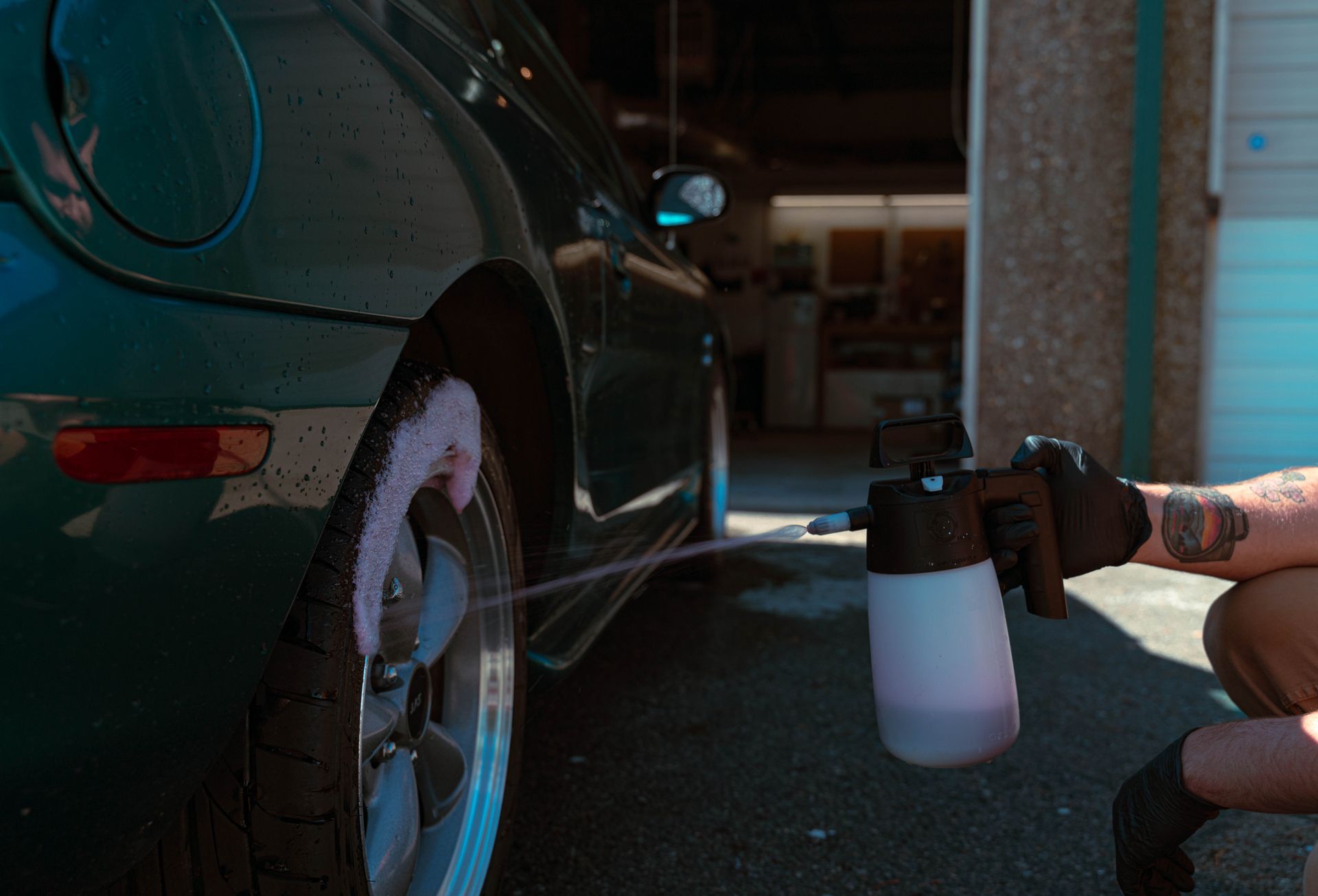 Person spray-washing a dark green car’s rear wheel in a garage.