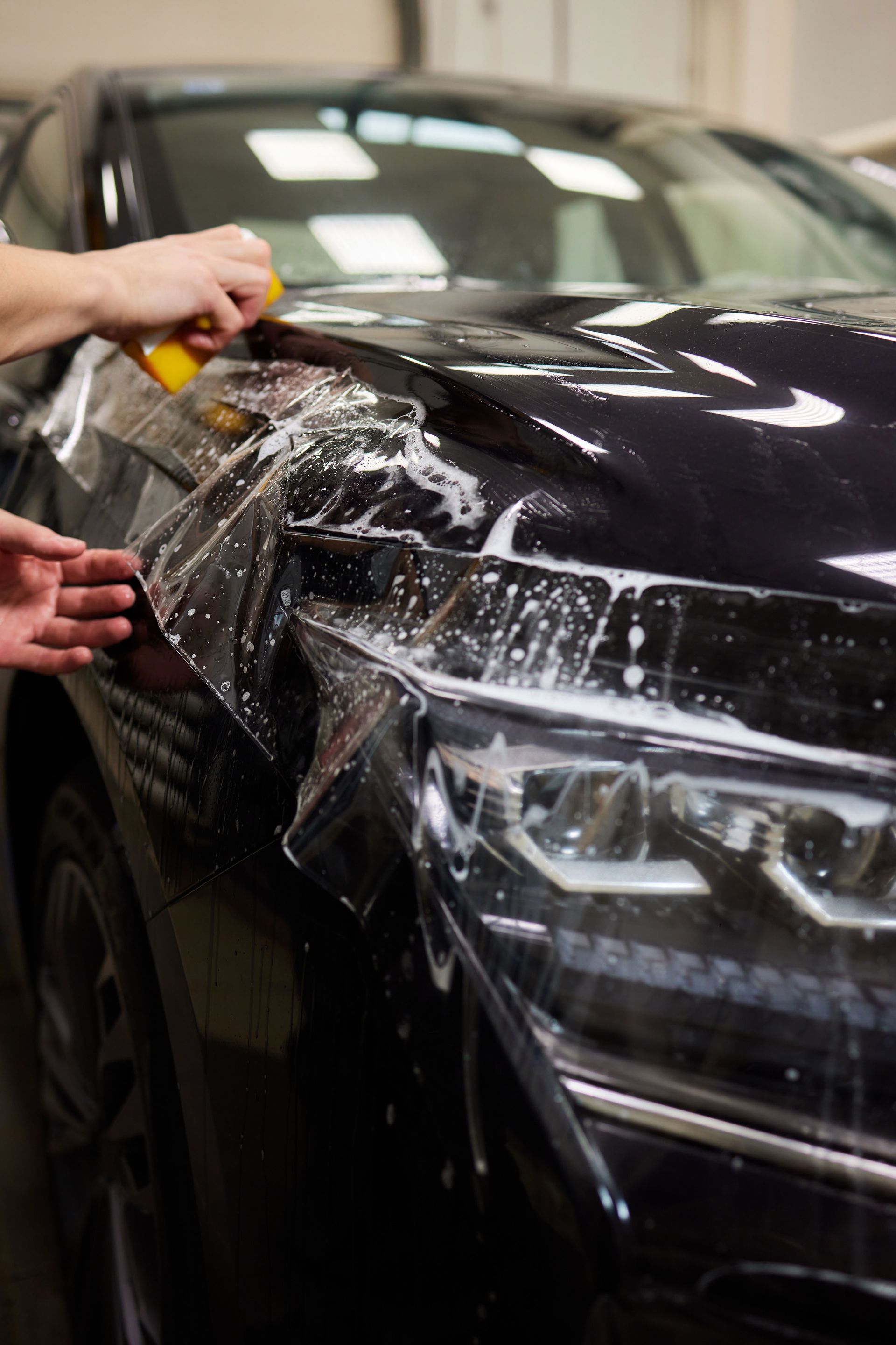 Hands washing a black car’s front fender and headlight with soap in a garage.