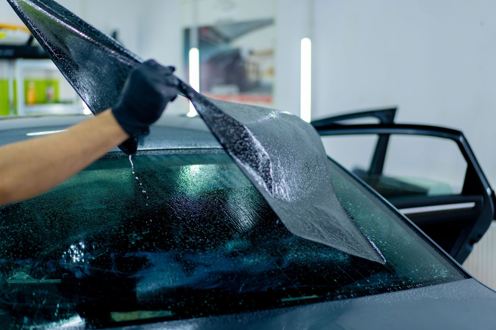 Gloved hands peeling shattered windshield film from a car in a repair shop