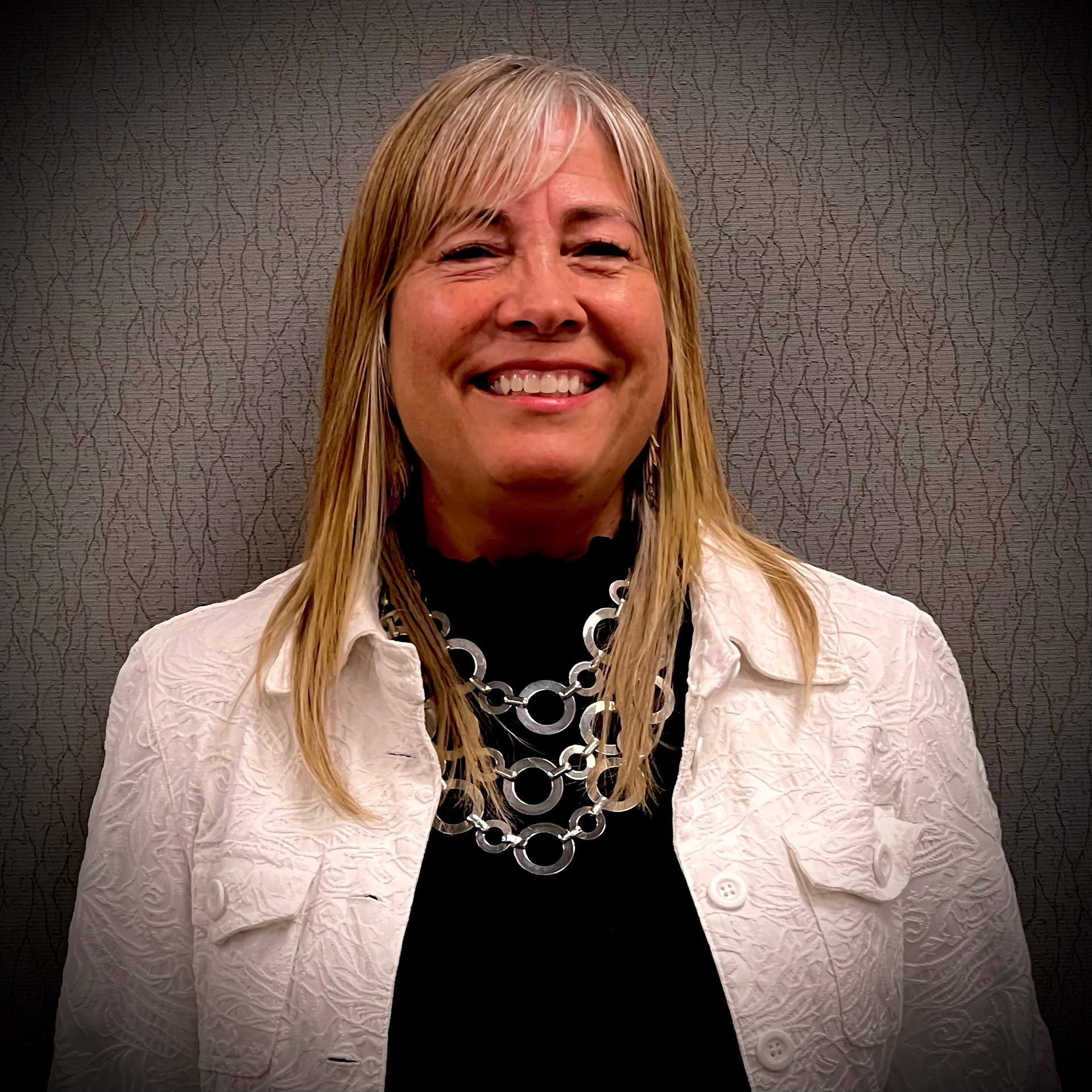 Smiling woman in a white jacket and silver necklace against a gray textured backdrop