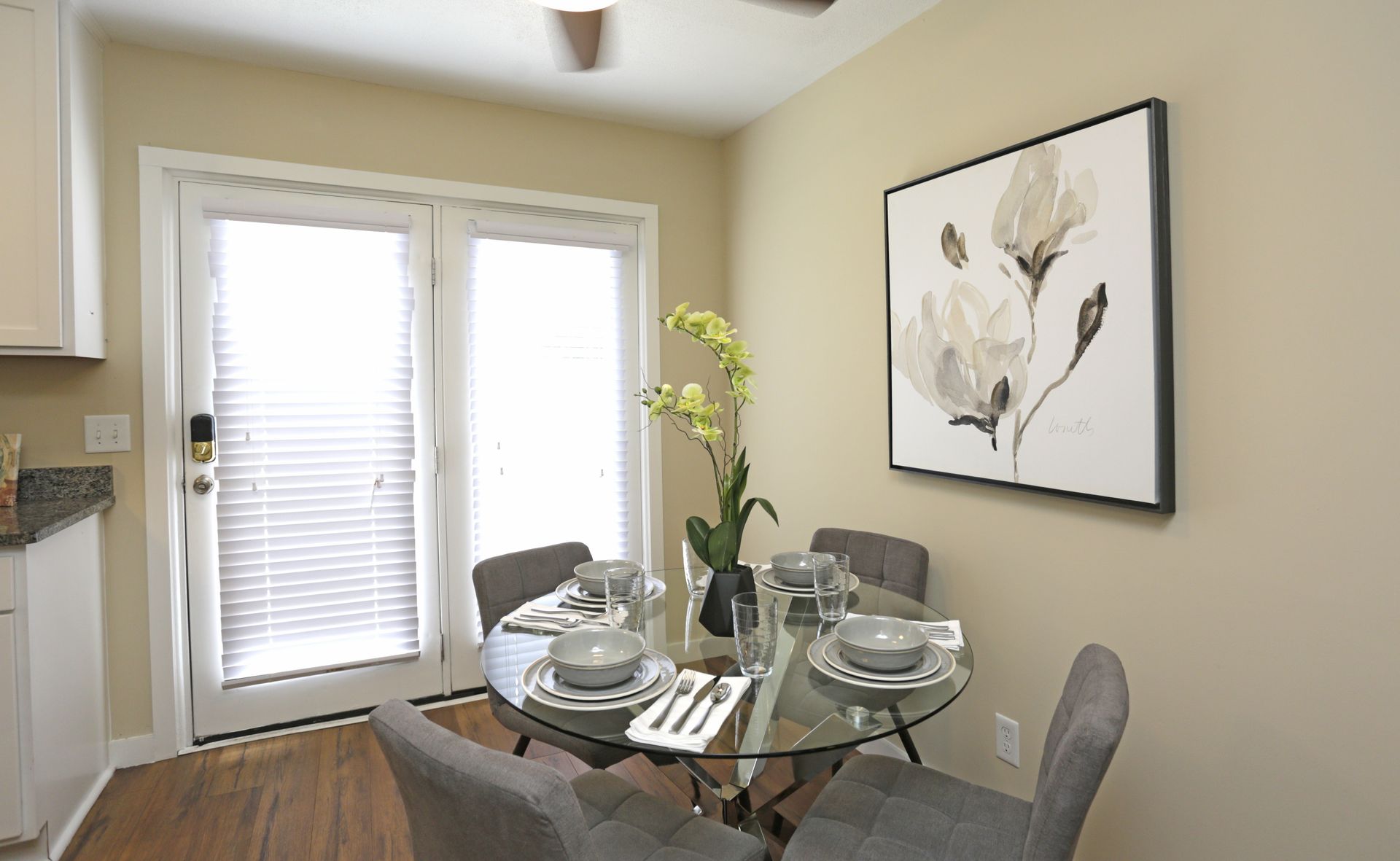 Dining area with glass table, four gray chairs, floral art, and French doors.